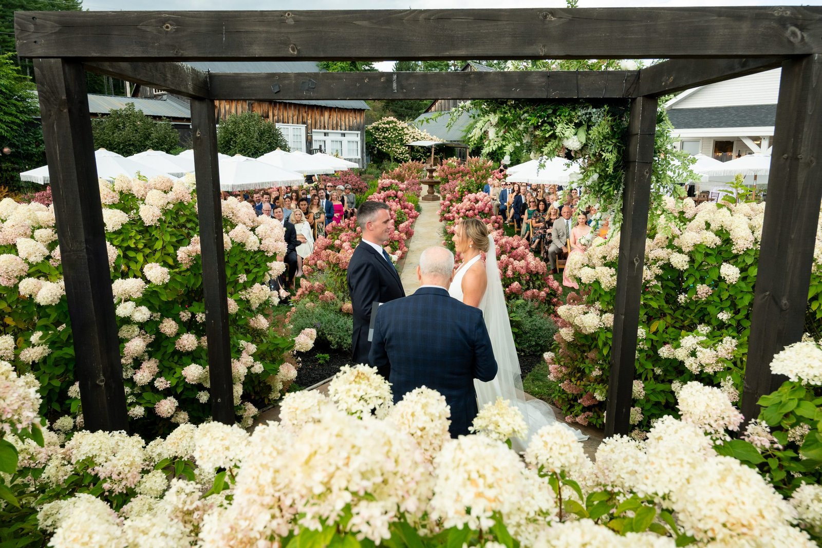 A bride and groom stand under a floral arch during their wedding ceremony, surrounded by lush, blooming flowers in soft whites and pinks.