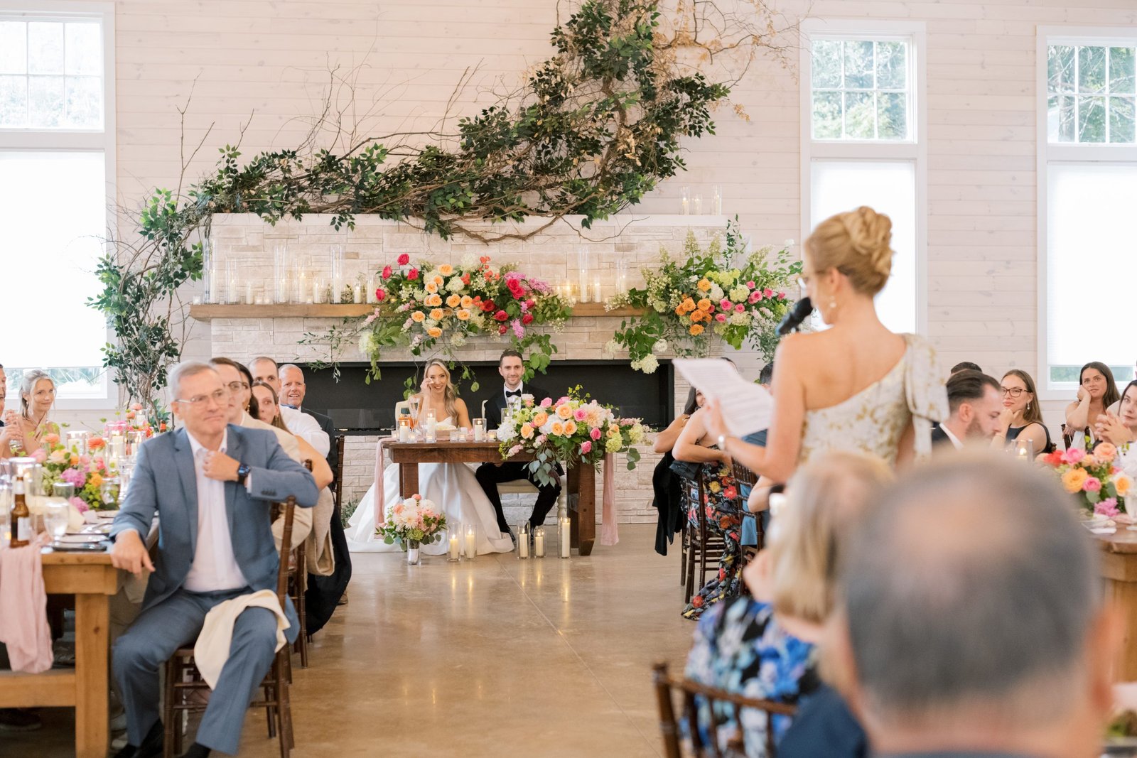 A wedding guest giving a speech during a reception with floral arrangements and a cozy fireplace in the background.
