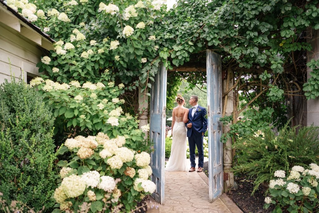 A bride and groom stand together in a charming garden doorway, framed by lush greenery and blooming white flowers.