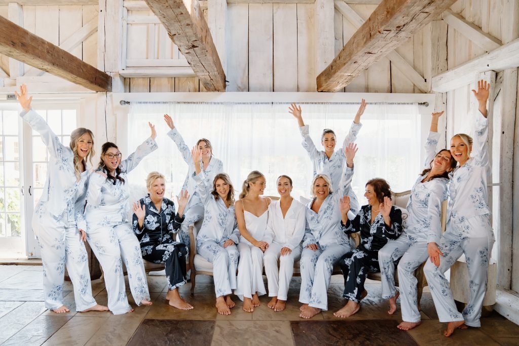 A joyful bridal party poses together in matching pajamas, with arms raised in celebration. The group of women, including the bride in the center, are seated and standing in a rustic indoor setting with natural light, radiating excitement and happiness
