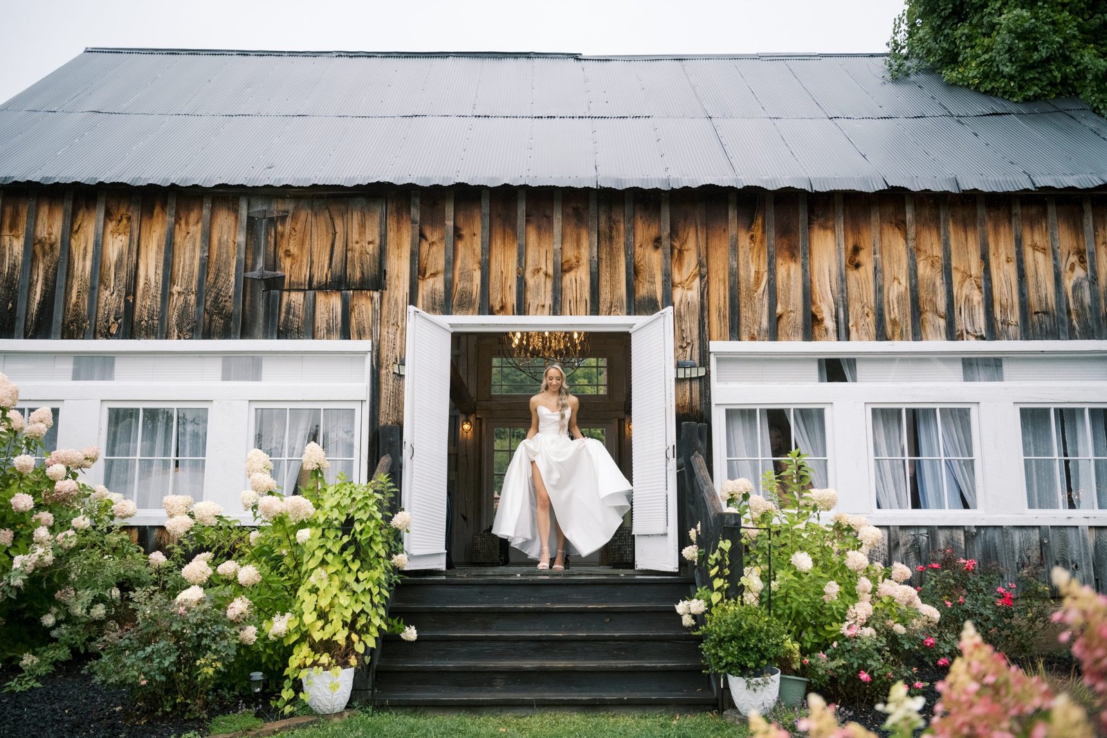 A person stands in the open doorway of a white building with a gray roof, surrounded by lush greenery and colorful flowers, with steps leading up to the entrance.