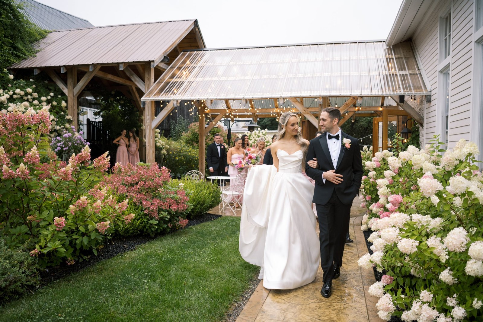 A couple dressed in wedding attire walks hand in hand down a garden path lined with blooming flowers, with guests gathered nearby and a rustic-style building in the background.