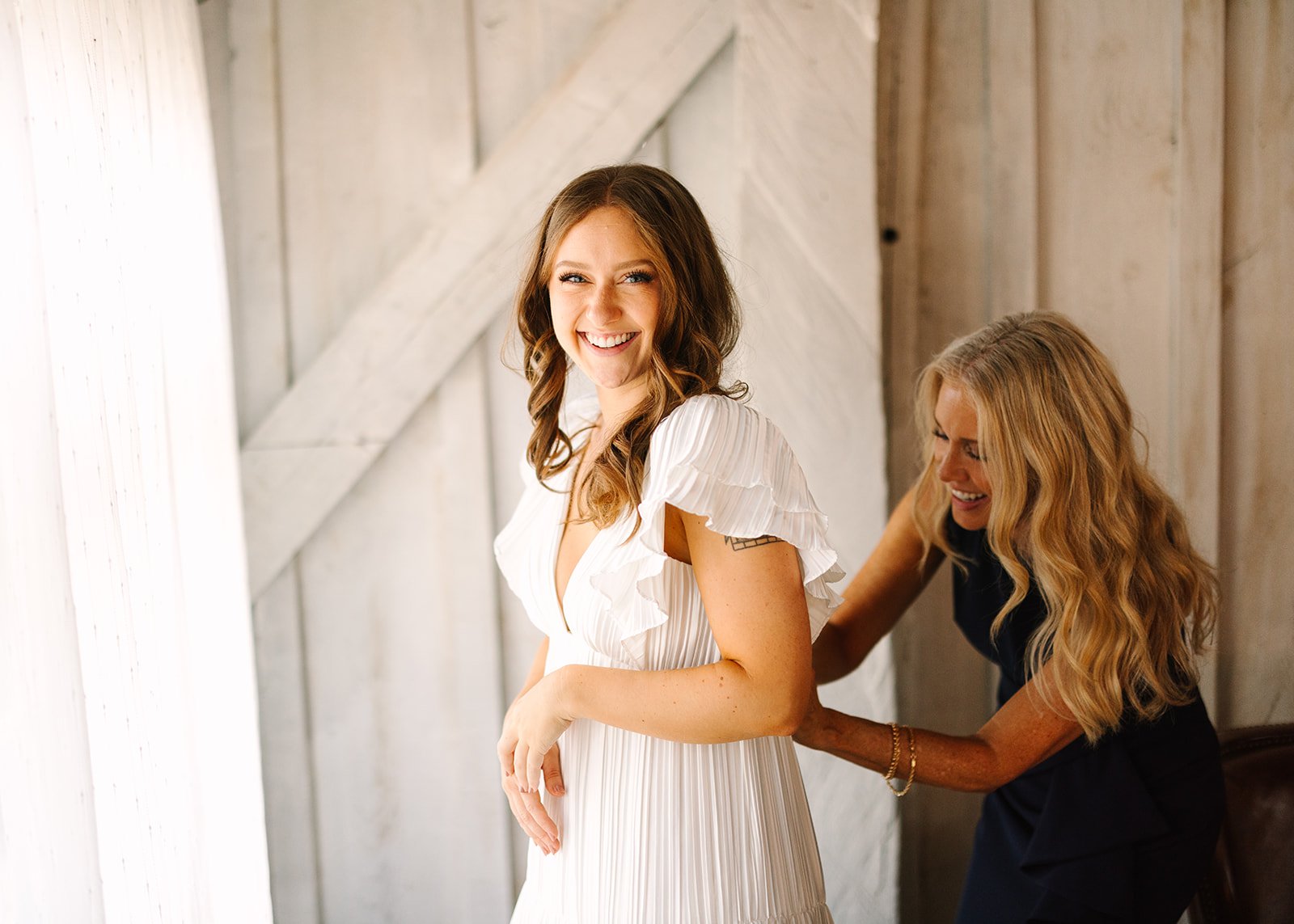 A smiling pregnant woman in a white dress stands indoors while another woman gently measures her baby bump with a measuring tape near a window.