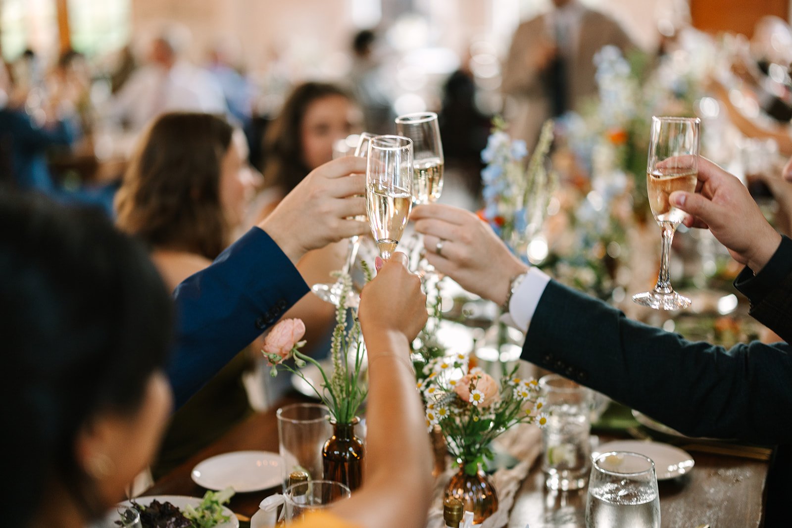 A group of people seated around a dining table raise their glasses in a toast, with plates of food and floral decorations visible, creating a warm and celebratory atmosphere.