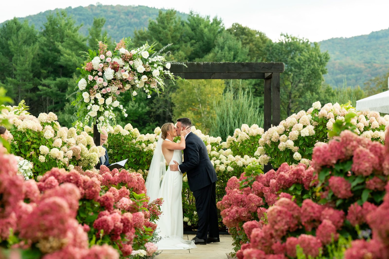 Bride and groom sharing a kiss under a floral arch, surrounded by hydrangeas and green hills, during their outdoor wedding.